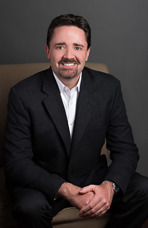 man sitting in chair inside with black suit smiling with hands crossed in front of gray wall, atlanta portraits, commercial portraits, business portrait photographers