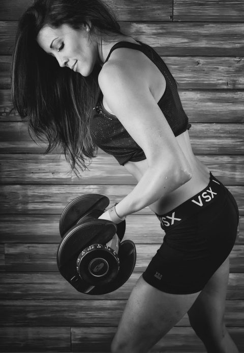 black and white woman in athletic attire standing in front of wooden wall lifting dumbbells and smiling, fitness blogger, postpartum fitness, workout clothes, fitness clothes, fitness attire, women's fitness, athletic photography, atlanta athletic photography, atlanta athletic photographer, athlete, athletic portraits, athletic portraiture, athletic portrait
